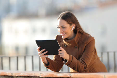 Happy woman wearing wireless earbuds in winter watching videos on tablet in a balconyの写真素材