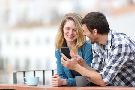 Happy adult couple talking about smart phone content standing outdoors in a balcony in a rural townの写真素材