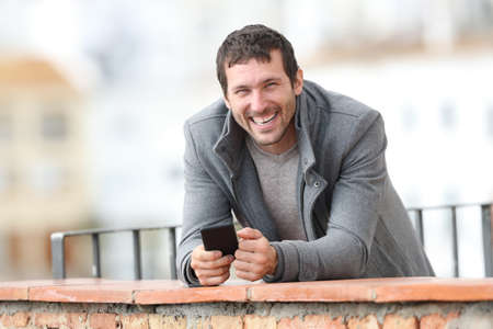 Happy adult man holding phone looks at camera in winter standing in a balcony outdoors in a rural townの写真素材