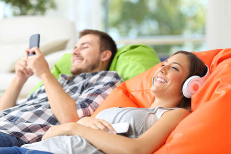 Happy couple enjoying using mobile phone and listening to music lying on colorful poufs at homeの写真素材