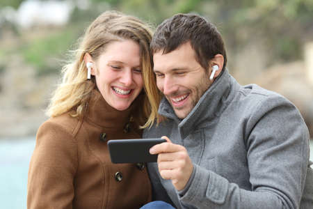 Happy couple watching videos on smart phone sharing earbuds on the beach in winterの写真素材