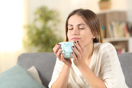 Relaxed woman smelling coffee from a mug sitting on a couch at home