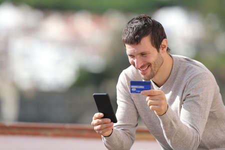 Happy man buying online with credit card and mobile phone sitting in a rural townの写真素材