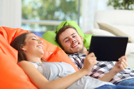 Happy couple lying on colorful poufs at home sharing tablet online contentの写真素材
