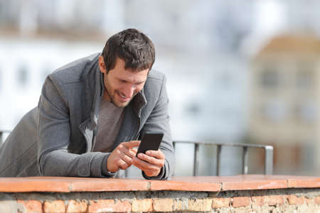 Happy man in winter using mobile phone standing in a balcony outdoors in a rural townの写真素材