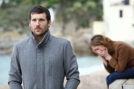 Sad wife complaining after break up sitting on the beach and angry husband leaving her in winterの写真素材