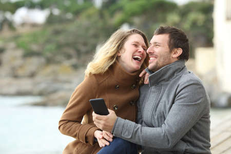 Happy couple of adults laughing loud about mobile phone content sitting on the beach in winterの写真素材
