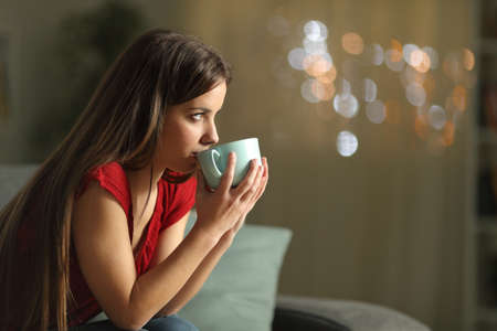 Side view portrait of a woman looking away drinking coffee sitting on a couch in the night at homeの写真素材