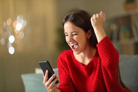 Excited woman in red checking mobile phone news sitting on a couch in the night at homeの写真素材