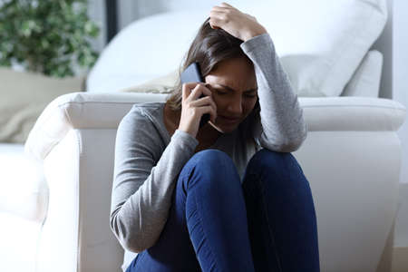 Sad woman complaining sitting on the floor talking on phone at homeの写真素材