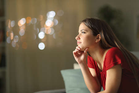 Profile of a pensive woman looking away sitting on a couch in the night at homeの写真素材