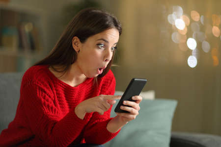 Surprised woman in red checking smart phone sitting on a couch in the night at homeの写真素材