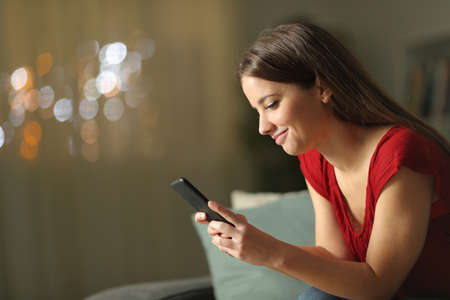 Profile of a woman in red using mobile phone sitting on a couch in the night at homeの写真素材