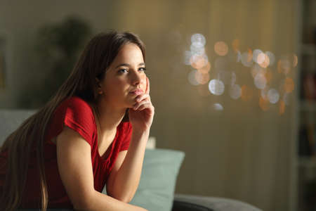 Pensive woman in red looking away sitting on a couch in the night at homeの写真素材