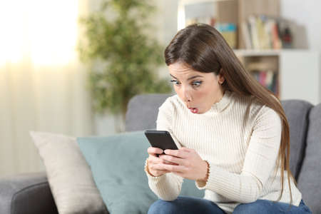 Surprised woman checks smart phone content sitting on a couch in the living room at homeの写真素材