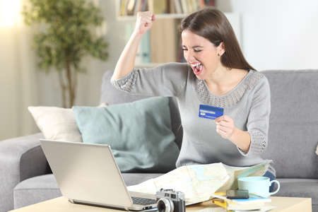 Excited tourist paying on-line with credit card planning vacation sitting on a couch in the living room at homeの写真素材