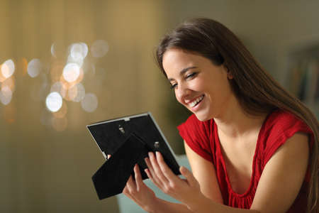 Happy woman remembering good times looking at picture frame sitting on a couch in the night at home