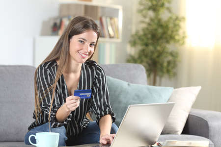 Happy consumer looking at camera holding credit card sitting on a couch in the living room at homeの写真素材