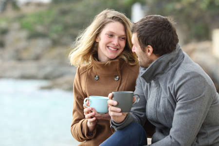 Happy couple of adults talking drinking coffee sitting outdoors in winter on the beachの写真素材