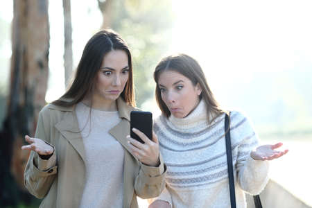 Front view of two perplexed women checking mobile phone content standing in a parkの写真素材