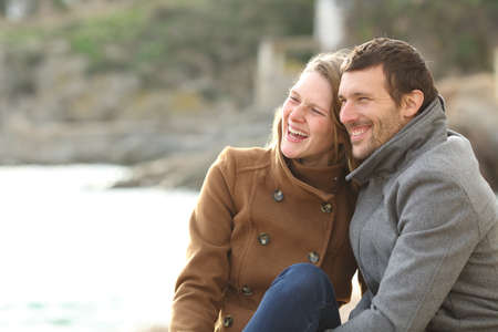 Adult couple dating and laughing loud sitting on the beach looking away in winter holidaysの写真素材