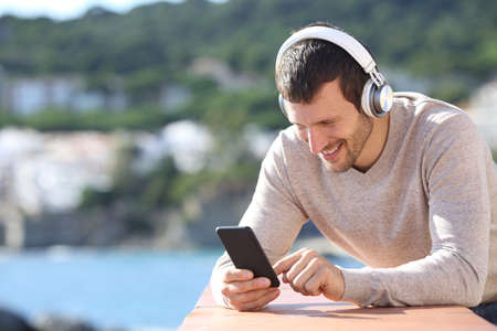 Happy adult man with headphones checking smart phone content in a balcony on the beachの写真素材