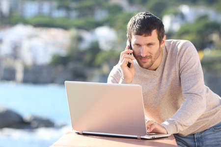 Serious man using a laptop and talking on mobile phone in a balcony on the beachの写真素材