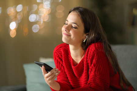 Woman wearing wireless earphones listening and feeling music from smart phone sitting on a couch in the night at homeの写真素材