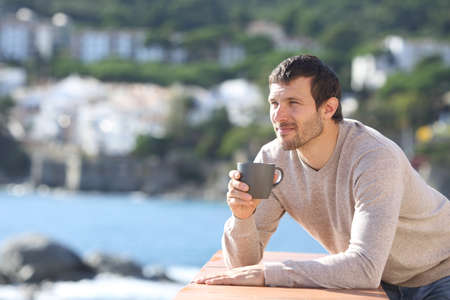 Serious man drinking coffee contemplating views in a balcony on the beach a sunny day