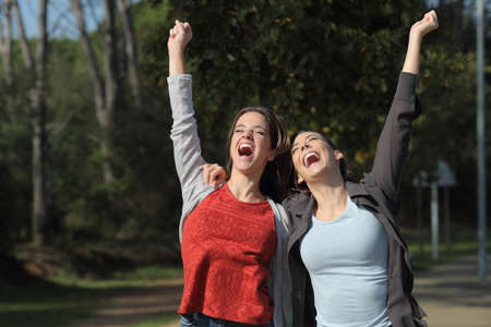 Front view portrait of two excited friends celebrating success raising arms in a park
