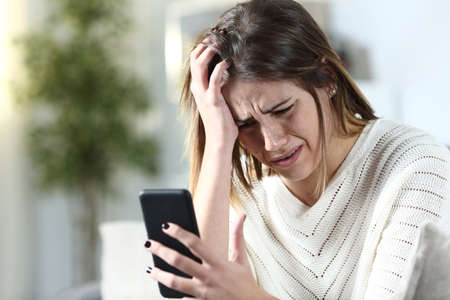 Sad girl complaining holding smart phone sitting on a couch in the living room at home