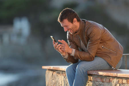 Happy adult man using mobile phone sitting on a wall of a balcony in a coast town