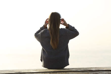 Back view of girl putting headphones sitting on a ledge outdoorsの写真素材