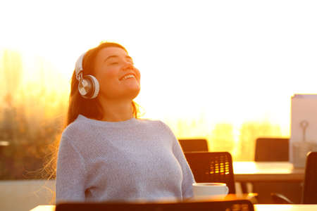 Happy girl relaxing listening to music wearing headphones sitting in a terrace at sunset
