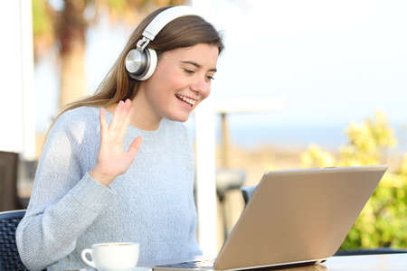 Happy girl greeting during videocall on laptop sitting in a coffee shop terrace on the beachの写真素材