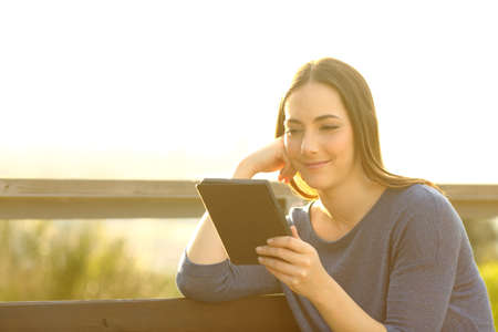 Woman reading an ebook sitting on a bench at sunset outdoors in city outskirtsの写真素材