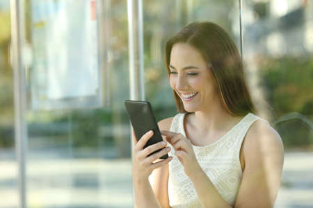 Happy young woman checking smart phone waiting for the bus to comeの写真素材