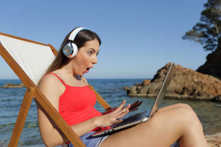 Amazed tourist with headphones looking shocked at her laptop on the beachの写真素材