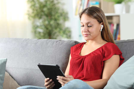 Woman wearing red shirt with wireless earbuds playing videos on a tablet sitting on a couchの写真素材