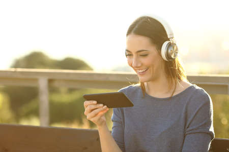 Happy woman watching videos with headset on her smart phone at the park at sunsetの写真素材