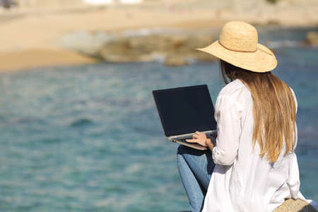 Back view of a woman wearing a pamela typing on her laptop with a sea background on the beachの写真素材