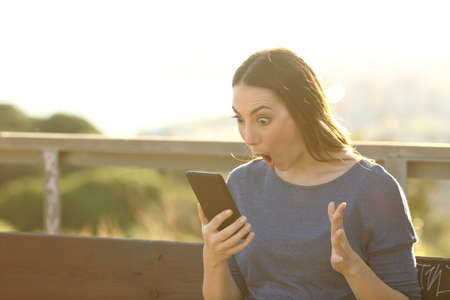 Surprised woman looking shocked at her smart phone screen on a park bench at sunsetの写真素材