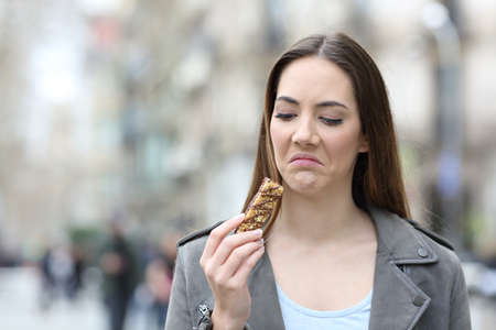 Front view portrait of a disgusted woman looking at snack cereal bar on a city streetの写真素材