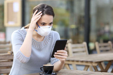 Worried woman with protective mask looking at smart phone reading news on a coffee shop terraceの写真素材