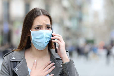 Portrait of a scared girl with hand on chest wearing protective mask calling doctor on smart phone on a city streetの写真素材