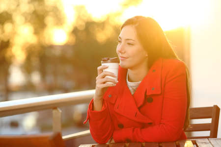 Pensive woman holding takeaway coffee cup at sunset sitting on a balconyの写真素材