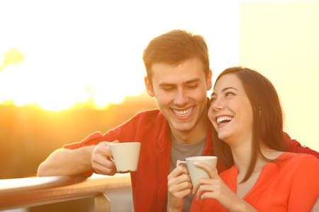 Happy couple cuddling holding coffee cups enjoying at sunset standing on a balcony at homeの写真素材