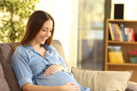 Happy pregnant woman expecting looking at belly sitting on a sofa in the living room at homeの写真素材