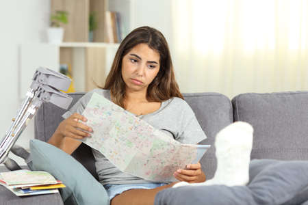 Sad disabled girl checks touristic map after canceling vacation sitting on a sofa in the living room at homeの写真素材
