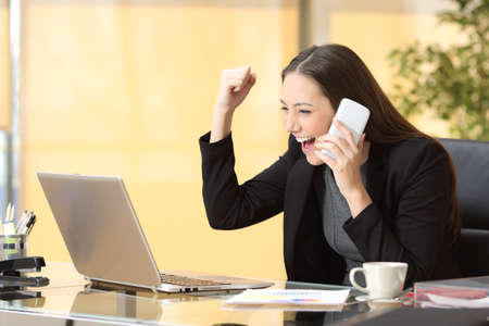 Executive woman talking on smart phone celebrating good news with laptop at the officeの写真素材